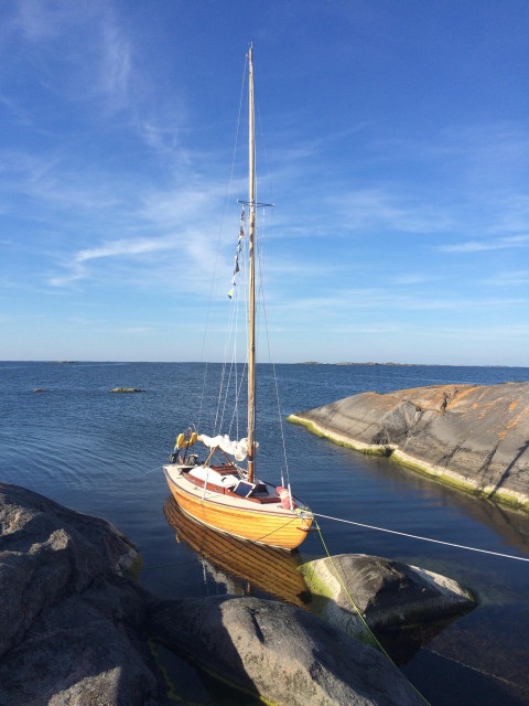 En folkbåt ligger föröjd mellan två klippor. Blå himmel med några vita molnstrimmor och bara horisonten bakom.
