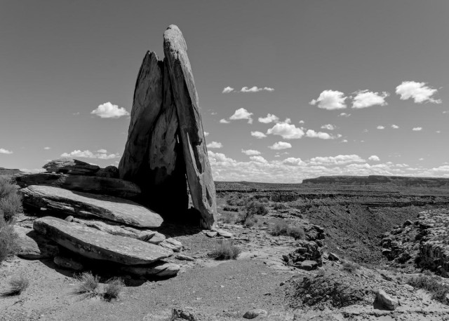 A black and white landscape photo of several very large flat rocks on the left and a canyon on the right. The sky has a few small puffy clouds on and above the distant horizon. Four of the rocks on the left are lying flat in a stair step pattern with the lowest one nearest the camera. Three of the large bus sized rocks are leaning together standing straight up on end looking a bit like a teepee.