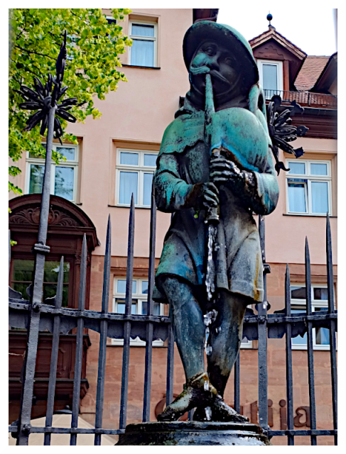 The image shows a statue of a person playing a musical instrument, possibly a flute. The statue stands in front of a wrought iron fence with vertical bars. Behind the statue is a building with several windows and a sloping roof. The statue is detailed and shows the figure wearing a hat and cape. On the left there is a tree with green leaves, partially visible. The statue appears to be made of metal, with some weathering visible on its surface.