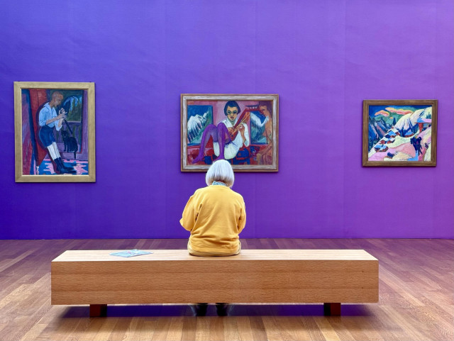 The Kirchner museum in Davos. Current exhibition is about colour. Image description: an art gallery with a purple wall featuring three paintings. A person wearing yellow with gray hair sits on a wooden bench, facing the artworks.