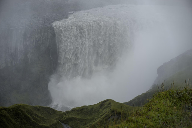 Dettifoss waterfall found in the Northeast of Iceland, described as one of the most powerful waterfalls in Europe. A steep 44meter drop, with very wide 100meters of water forcefully crashing down, kicking up so much spray that the area below is blanketed in a constant grey mist. Across the other side of the waterfall can be seen some people stood on the rocky landscape, their size very small in comparison to the waterfall. On the side where this photo was taken, is some mossy vegetation covering the jagged and downward sloping terrain.