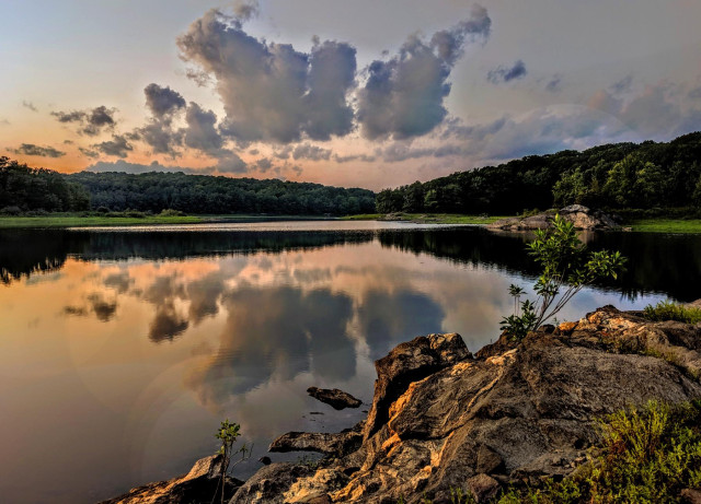 We are standing next to a big rock on the shore of a large lake looking across the lake. The opposite shore is a wide strip of grassy looking plants and then forest. The evening sky is a very pale blue, with a line of large and small clouds drifting past above the horizon.