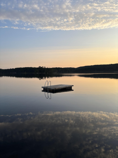 A tranquil lake scene with a wooden swimming platform on calm water, reflecting clouds and a colorful sky at sunset. Trees lines in the horizon. 