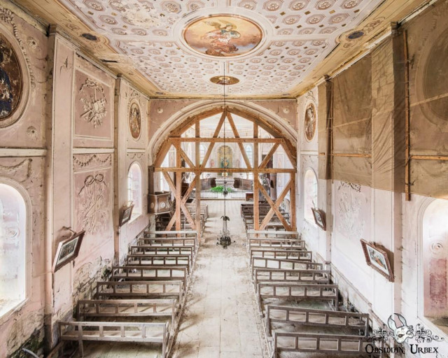 An abandoned, ornate church interior with peeling pink paint and rows of broken pews. A wooden scaffold stands before the altar, under a beautifully frescoed ceiling