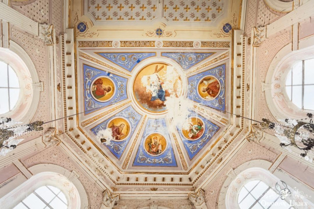 A beautifully painted domed ceiling inside an old chapel features a central religious scene and surrounding saints. Ornate patterns in blue, gold, and pink adorn the ceiling.