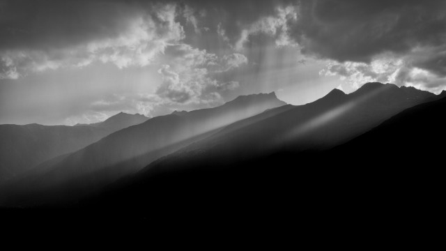 A monochrome landscape with stark contrasts showing mountain silhouettes against a cloudy sky, with rays of light filtering through the clouds, creating a moody atmosphere.