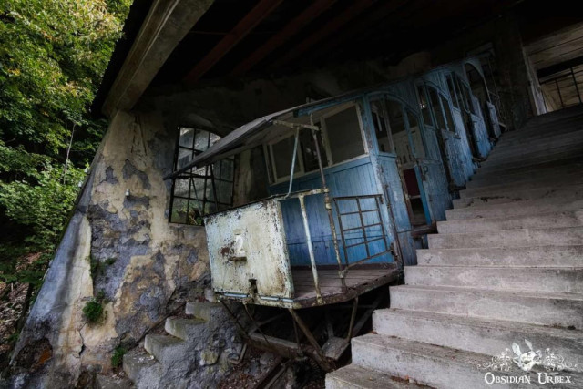 An abandoned blue funicular car at an old alpine railway station, featuring crumbling walls and concrete steps.