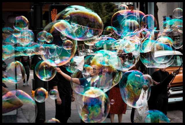 A street photo of a cluster of people... Two of whom appear to be waving their arms in the air, releasing a cloud of giant soap bubbles on a street corner. You can see the joyful miscreants, arms raised, through some of the bubbles they have released. 

Taken at a Sunday Streets event, where a dozen blocks of Valencia Street were closed to traffic and opened to vendors, entertainers, and people.