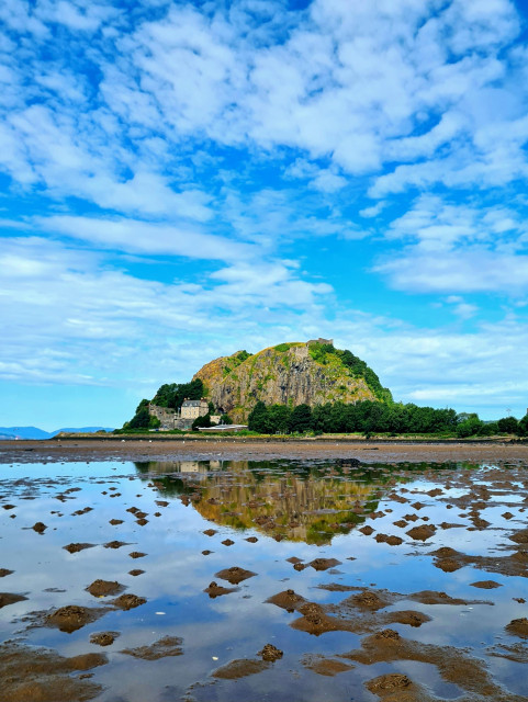 A volcanic plug with fortificationa on in reflected in a still water pool left by the receding tide.