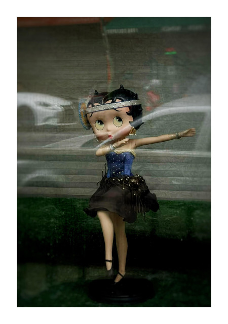 Photo of a Betty Boop figurine, one of several Betty Boops in a quirky window display in an apartment in Red Hook, Brooklyn. This one has been posed in a dancing position almost like a dap, with one arm extended and the other bent at the elbow, and with one leg raised. She’s wearing what looks like a 1920s flapper outfit: short black skirt, blue corset studded with pearls, silver bracelets, gold earrings, and a silver jewel-studded headband with a large blue-rimmed disc to one side. The figurine stands on a surface with a little step behind, all covered in green felt. The window glass reflects some cars parked at the curb and a storefront across the street.