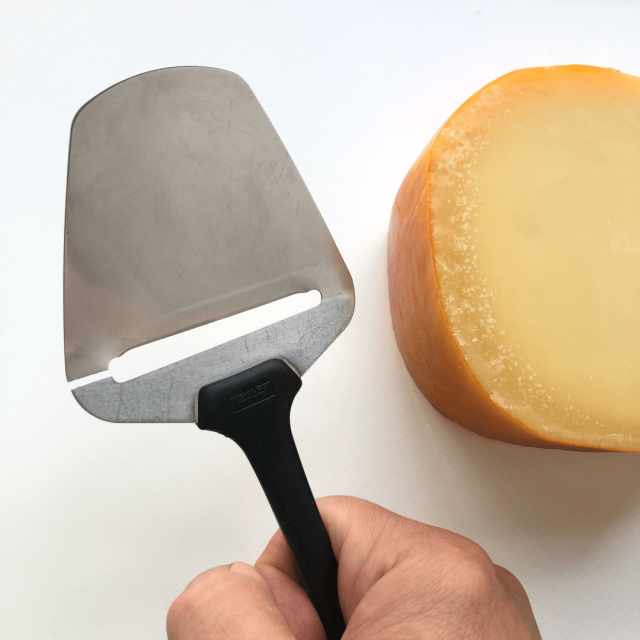 Against a white background, a hand holds a broken cheese slicer (kaasschaaf). On the right of the frame a block of cheese is visible.