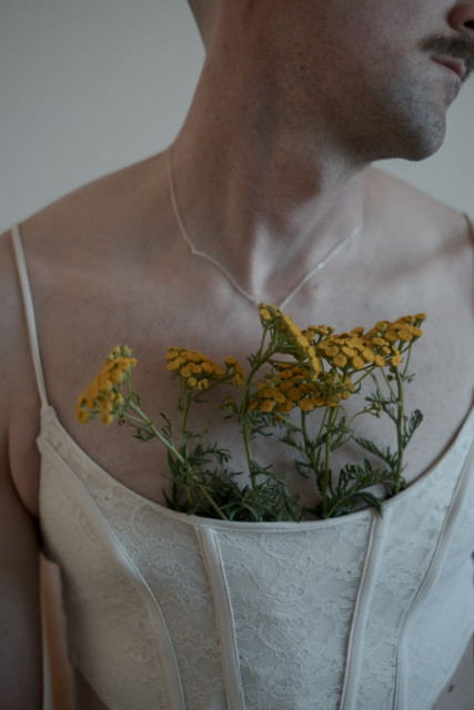 Close-up portrait from chin to waist showing a person with facial hair wearing a white lace corset. Bright yellow wildflowers are tucked into the top of the corset. A thin necklace rests on bare skin against a soft neutral background.