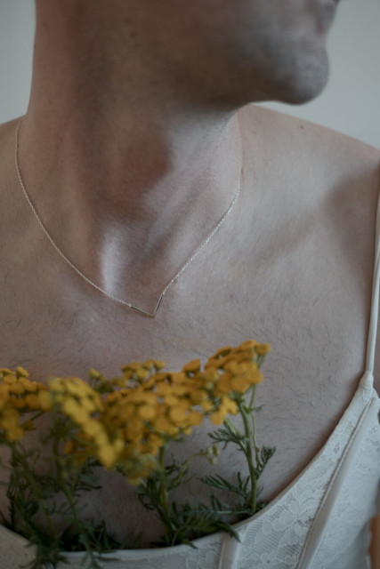 Close-up portrait of a person's neck and upper chest, showing chin with stubble, wearing a delicate silver V-shaped pendant necklace and a white lace top. Bright yellow flowers with green stems are held against the chest. Soft, warm lighting against a plain off-white background.