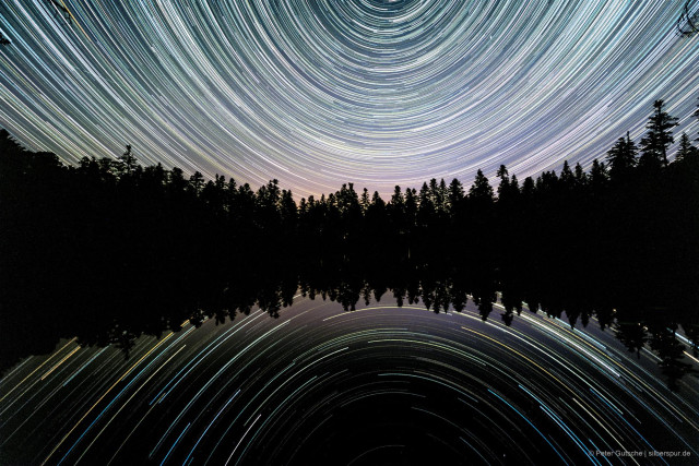Night sky over a calm lake: Star trails are reflected symmetrically in the water. Because the camera was pointed directly north and the horizon with the tree line on the opposite shore was placed in the center, the star trails form two concavely curved circular arcs that are mirrored in the lake.