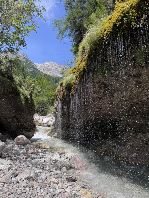 Ein Gebirgsbach fließt durch eine Engstelle. Rechts im Bild rinnt und tropft Wasser wie ein Vorhang von einem unten schon mannshoch unterspülten Abhang mit Bäumen. Im Hintergrund hohe Berge und blauer Sommerhimmel. 
