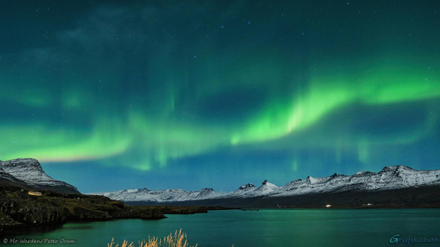 A colour photo of a night sky above a body of water. The shores of the fjord are lined on both sides by ranges of snow-capped mountains, lit by moonlight from behind the viewpoint. The foreground is out of shot except for an area of yellow grass heads. On the left shore is a lit building. The sky is full of stars including part of the Ursa Major asterism. From behind and above the mountains on the left and stretching across the photo are bands of green aurora. Some brighter bars are standing in the streams, and the colours are reflected in the water surface. On the left at the bottom of a strong arc of aurora is a pink edge below the green. This is the nitrogen fringe.