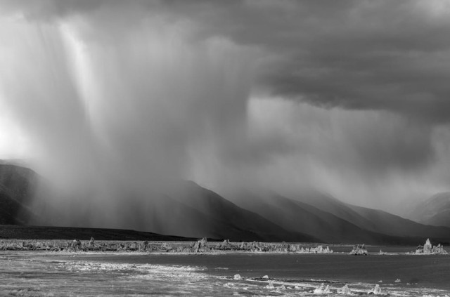 A black and white landscape of heavy rain over the front range of tall mountains. The rain is pouring heavily in a cylindrical shape in one area of the mountains on the left. Lighter rain is seen on the right behind the area of dense heavy rain. Part of a lake is seen in the foreground on the right.It's a wedge shape coming in from the right and forming a point on the left. Form the point on the left a sliver on land extends across the image to the right. It has small spike like formations called tufa towers. 