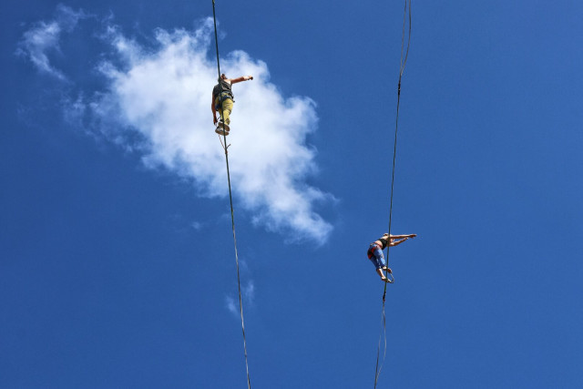 Two people suspended on vertical ropes against a vibrant blue sky. One person, wearing a camouflage top and light trousers, has one arm outstretched for balance. The other person, in a blue outfit, also extends their arms. Both wear safety harnesses. A single white cloud floats between them.