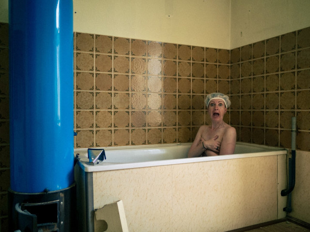 Eine Badewanne vor braun gemusterten Kacheln, daneben ein dickes blaues Rohr.
In der Wanne sitzt eine Frau mit Duschhaube und guckt sehr erschrocken.