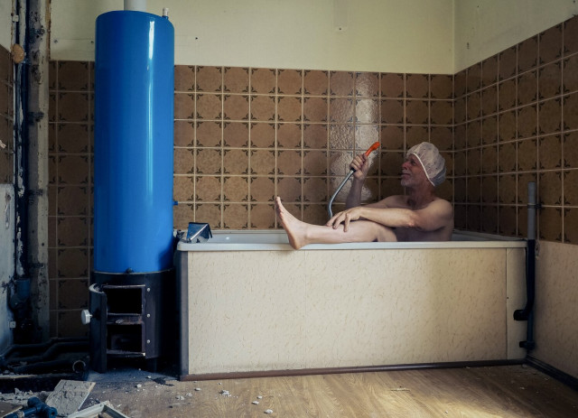 Eine Badewanne vor braun gemusterten Kacheln, daneben ein dickes blaues Rohr.
In der Wanne sitzt ein Mann mit Duschhaube und duscht sich - trocken - das Gesicht, wo bei sein Bein auf dem Wannenrand liegt.