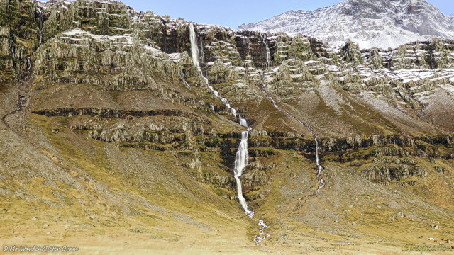 A colour photo of a brown rock mountainside with a waterfall cascading down it. Above and to the right is the mountain peak, covered in snow and ice. A small amount of clear sky can be seen. The rock face is layered volcanic rock with scree slopes at intervals down its drop. The lower slopes are almost completely obscured by angled scree with vegetation. Near the centre of the shot is a white water horsetail falls which quickly encounters the rocks. Below that point is a series of about ten basalt steps, and the water drops over each of them, finally disappearing into the lower scree. Several smaller cascades are on either side.