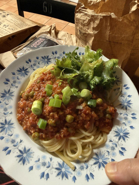 A close-up view of a hand holding a floral-patterned plate filled with spaghetti topped with a rich tomato ragù, green peas, chopped spring onions, and a side of fresh green lettuce. Takeout paper bags and packaged food sit in the background.