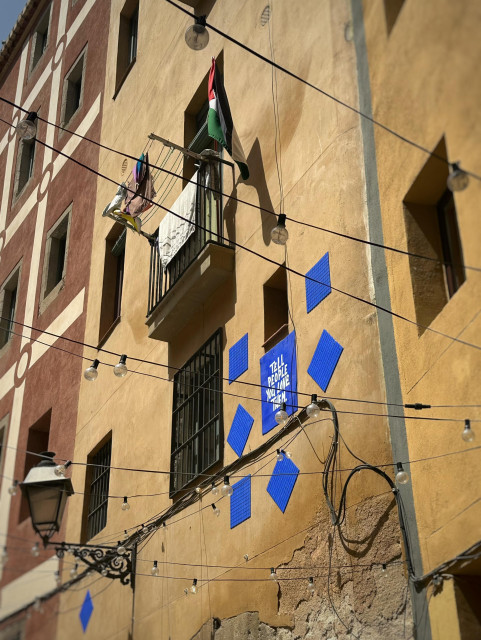 Detalj mot en balkong med hängande tvätt, en Palestinaflagga och en blå plakett som det står Tell people you love them på och med andra blå kvadrater bredvid.