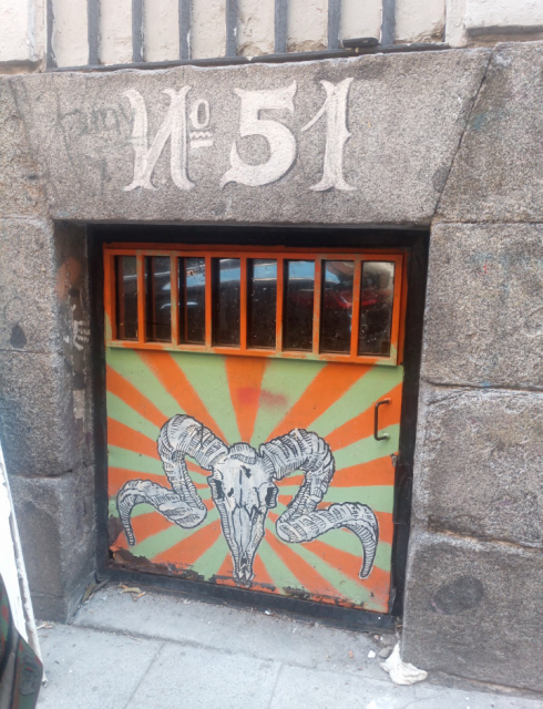 A roughly metre square aperture in a large stone wall of a building that's floor level has a huge 51 above and a metal door with mirrored grate at the top. It's painted orange and green with a goat's skull in the centre.