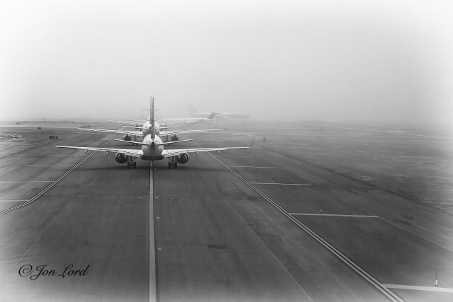 This is a black and white photo in landscape format of a busy airport taxiway surrounded by fog with a long line of aircraft waiting to takeoff. JFK, New York (2011).

One third of the way to the right of the left edge of the photo is a long solid line stretching along the centre of the taxiway and away from the camera. A short distance to the right of the centre line (about ten cm) is a line of lights inset into the tarmac, spaced every few metres. About ten metres to the left and right of the centre line are two parallel solid lines marking the edge of the taxiway, one either side, though the tarmac continues for a distance of another five or ten metres beyond. Further to the right is a wide grass area before a small portion of a parallel runway is in view. Surrounding this scene is a bright, white, morning fog with a visibility of no more than five hundred metres. In front of the camera is a row of five aircraft on the taxiway waiting for their turn to take off. The closest aircraft is about ten metres from the camera, is a Boeing 737 narrow body, shorthaul aircraft with two jet engines slung under the wings and small winglets on the wingtips. Beyond the queue of aircraft is a further larger aircraft, about five hundred metres away, at right angles to the taxiway and is on the runway and starting its takeoff roll, this aircraft is partially obscured by the fog. Above and beyond is filled with a large body of bright, morning fog.