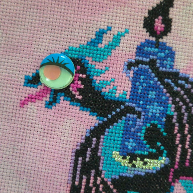A cross stitch of a black dragon wrapped around a candlestick with a black flame.  There's a large, googly eye with eyelashes placed over the eye of the dragon, making it look ridiculous. 