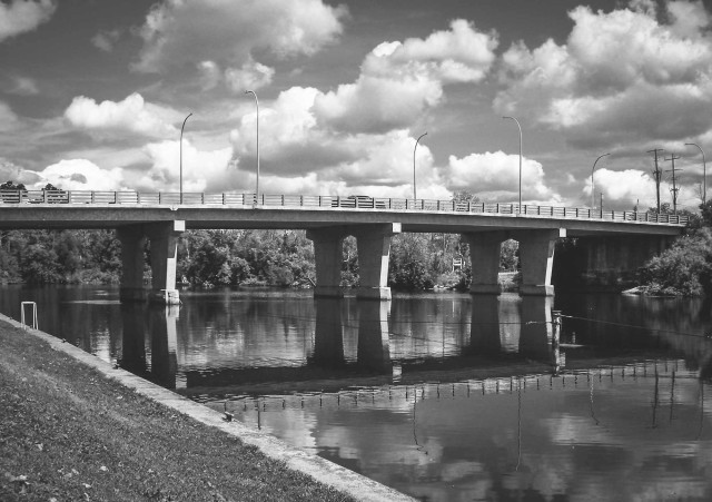 A road bridge crosses the calm water of a river. Large white clouds fill the sky on this sunny summers day. This is a black and white image.