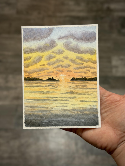 Hand holding a sunset painting with lots of dark shadowed clouds on the top with reflections in the water and some waves on the grey rocky shoreline in the foreground. Towards the middle there are some trees in silhouette. 
