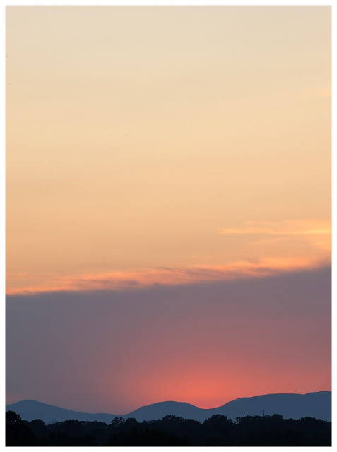 A hazy sunset behind distant hills, with a red-orange glow near the horizon and soft blue and pink tones in the sky above darkened silhouettes of trees and mountains.