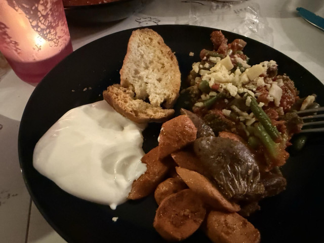 Close-up of a black dinner plate holding a serving of sliced cooked sausages, a slice of crusty bread, a dollop of white cream sauce, and vegetables in a tomato sauce topped with grated cheese, illuminated by a glowing pink candle nearby.