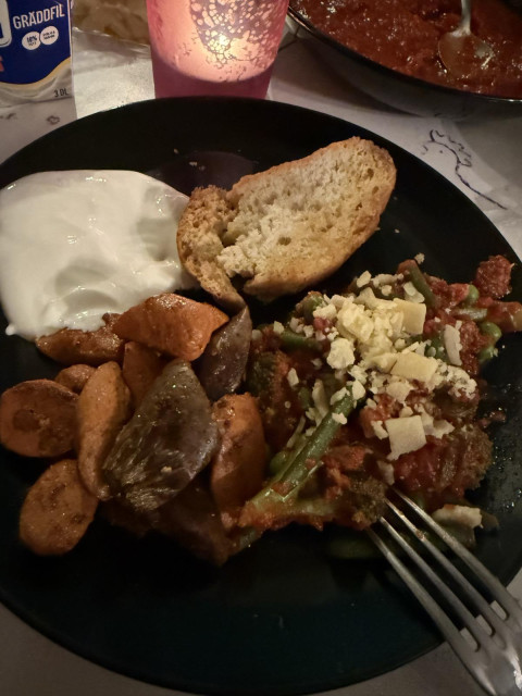 A top-down close-up of a dinner plate loaded with sliced sausages, roasted vegetables, a slice of bread, sour cream or yogurt, and a vegetable tomato ragù topped with grated cheese, with a fork resting on the side.
