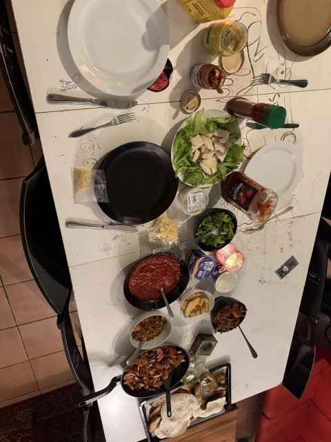 Top-down overhead view of a large dinner table spread. The table is filled with various dishes including sliced sausages in a frying pan, red tomato sauce, side salads, bread, condiments, and several place settings on a white table marked with faint drawings.