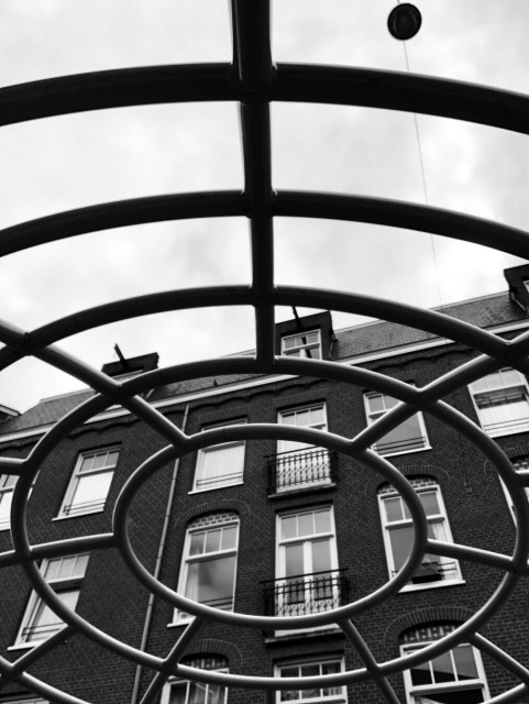 Looking upward from the inside of a playground set of bars towards a typical Amsterdam block of flats against a cloudy sky.