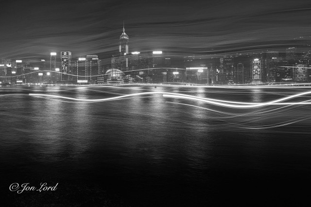 This is a black and white nighttime cityscape, long exposure photo of a body of water with the light trails of passing boats and a high-rise waterfront cityscape in the background. Victoria Harbour, Hong Kong (2018).

The dark harbour water rises from the base of the photo upwards to a little above the half way up level. The water at the base is pitch black with it becoming an undulating silver tone from the reflection of the bright lights of the distant, about one to two km away, high-rise waterfront cityscape. From the quarter up level to the top of the photo are a series of random wavy lines crossing the image horizontally from left to right, light trails caused by passing boats and a long camera exposure. Around the centre section of the photo, the lines are much brighter and clearly defined, with the lines towards the top having a dimmer less distinct, almost mist like appearance. Beyond, is the city's waterfront lined with multiple, brightly lit skyscrapers, many with large and illuminated signs on their roofs. Some with lighting in the shape of Christmas decorations stretching the height of those buildings, one with 'Seasons Greetings' in large neon lettering near the top. The rapidly rising terrain behind the waterfront is obscured by the misty light trails towards the top of the image. 