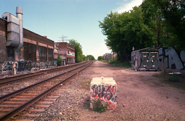 A graffiti clad concrete block stands next to two sets of parallel railway tracks behind graffitied industrial buildings. 