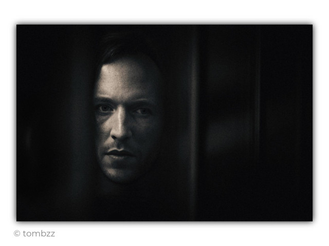 A black-and-white analog portrait of a man framed in a very tight, dark composition. His face emerges from deep shadow on the left side of the photograph, while the rest of the image is occupied by dark vertical bands resembling narrow openings, sections of bars, or parts of a heavy structure separating the subject from the viewer. It is unclear whether he is standing behind a door, behind some kind of screen, or simply within a narrow gap. This ambiguity creates tension and a sense of confinement.
The light falls selectively across his forehead, eyes, nose, and lips, leaving his cheeks and neck in shadow. The most important element is his gaze, directed straight into the lens. It is still, focused, and difficult to interpret: alert, cold, and unsettlingly calm at the same time. His face reveals no single clear emotion, making him appear like a character from a psychological thriller, someone who is watching or perhaps being watched himself.
The visual style recalls the dark psychological worlds of Ryan Murphy’s productions, where elegant imagery is often combined with unease, mystery, and hidden tension. The vertical divisions strengthen the cinematic quality of the portrait, while the grain, muted contrast, and deep blacks give the photograph a raw, claustrophobic atmosphere. The entire image is a deliberate construction built around ambiguity, confinement, and direct eye contact.