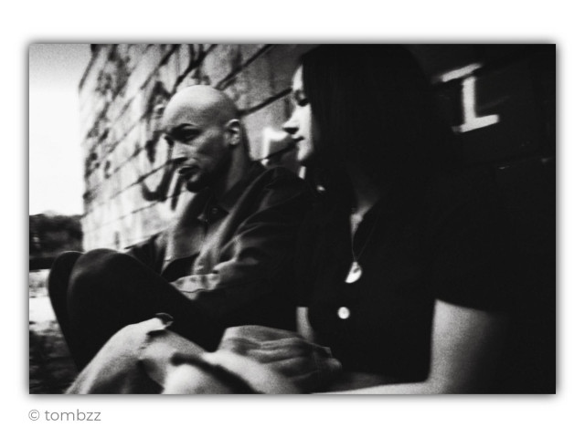 A black-and-white analog photograph of a young couple sitting on the ground against a brick wall covered with graffiti. Both lean against the wall and look somewhere beyond the frame, as though watching something in the distance or simply sharing a moment of silence.
The man has a shaved head, short facial hair, and heavy dark eye makeup that gives his face a raw, slightly theatrical expression. He is dressed in a dark jacket and trousers, sitting with his knees drawn up and his body leaning slightly forward. His gaze is focused and serious.
The woman sits closer to the camera. She has long, straight dark hair, and her pale face is partly immersed in shadow. She wears a dark short-sleeved top and a light-coloured skirt or fabric draped across her knees. A small pendant around her neck catches the light. Her profile appears calm, thoughtful, and slightly distant.
The frame is softly blurred, grainy, and strongly contrasted. Their dark clothing almost merges with the shadows, while the brick wall and fragments of graffiti create a rough urban background. The photograph strongly evokes dark wave aesthetics and alternative music culture. It carries a sense of melancholy, distance, and quiet rebellion, like a still from a low-budget music video from the 1980s or 1990s.