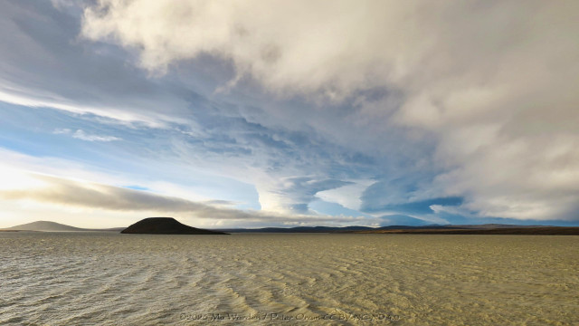 A colour photo of a rippled reservoir of pale brown water. On the horizon are some low mountains and near the centre is a cloud structure that's funnel-shaped. The top of the column widens up to a cloud bank overhead, with small spirals of cloud below it. In the foreground is a long curve of angry-looking cloud, which sweeps from the top left of the shot in an arc down to the lower right, then is flattened just above the water surface. It looks threatening and gives a sense of foreboding.