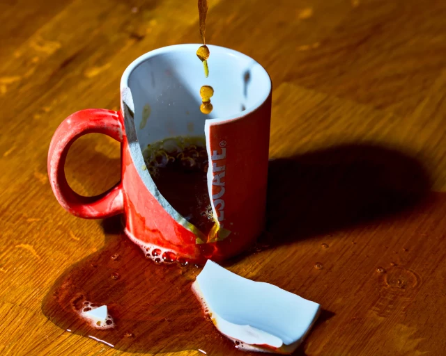 Eine rote Espressotasse steht auf einem Tisch. Ein großes, V-förmiges Stück ist aus der Tasse herausgebrochen und liegt davor. Von oben tröpfelt noch Kaffee nach, während sich eine Kaffeepfütze auf dem Tisch ausbreitet. 