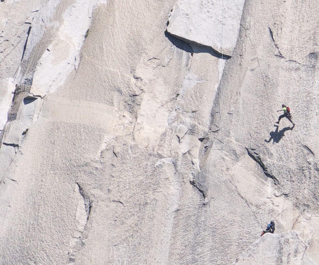A color landscape photo of a gray granite rock face with two climbers. One climber is spotting a second climber about thirty feet above them as they attempt to swing from the far right side of the image to the far left side of the image where they will grab hold of a crack in the rock face so they can continue their climb up the mountain. The climber attempting the swing from the right, and who is suspended from a rope above them, has a yellow-green shirt on, a small backpack, and is making a very large step to gain the momentum necessary to swing across the rock face to the target crack.