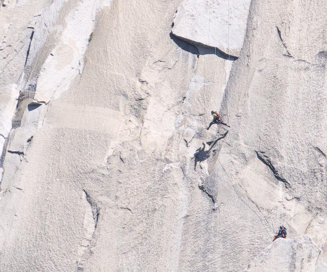 A color landscape photo of a gray granite rock face with two climbers. One climber is spotting a second climber about thirty feet above them as they attempt to swing from the far right side of the image to the far left side of the image where they will grab hold of a crack in the rock face so they can continue their climb up the mountain. The climber attempting the swing from the right, and who is suspended from a rope above them, is now at the bottom of the swing and is making very large strides in their attempt to swing far enough to grab the crack on the left side of the frame.