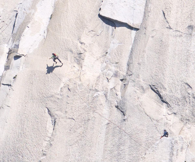A color landscape photo of a gray granite rock face with two climbers. One climber is spotting a second climber about thirty feet above them as they attempt to swing from the far right side of the image to the far left side of the image where they will grab hold of a crack in the rock face so they can continue their climb up the mountain. The climber attempting the swing from the right, and who is suspended from a rope above them, is now nearing the crack on the left and is just starting to reach out to insert their fingers into the crack. They are still about ten feet away, however.