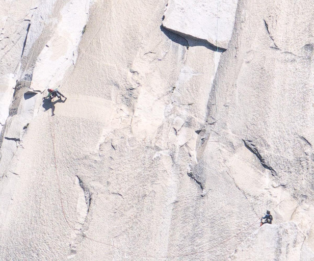 A color landscape photo of a gray granite rock face with two climbers. One climber is spotting a second climber about thirty feet above them as they attempt to swing from the far right side of the image to the far left side of the image where they will grab hold of a crack in the rock face so they can continue their climb up the mountain. The climber attempting the swing from the right, and who is suspended from a rope above them, has now reached the crack on the far left and appears to have obtained a hold. They are lying horizontal on the rock face and have yet to pull themselves over the crack.