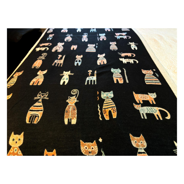 Photo of a bedspread with multiple stylized cats of different designs on a black background, viewed so that the bedspread recedes toward the far end of the bed at top frame. 