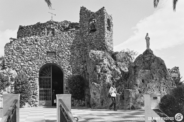 Black and white photograph of Ermita de la Virgen de la Peña with a bell tower, surrounded by rocky terrain. A statue stands on a rock beside the entrance, while a person walks in front of the church, framed by palm trees and a checkered path.
