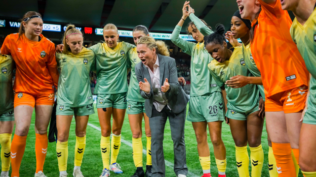 Vancouver Rise FC players in green-and-yellow uniforms and orange goalkeeper kits gather in a celebratory huddle around their coach, who leans forward clapping and shouting on the pitch inside a stadium.
